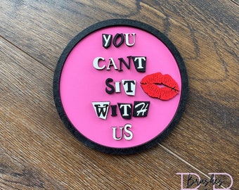 以下が含まれることがあります： 黒い縁取りのあるピンクの丸い看板に、白い文字で「YOU CANT SIT WITH US」と書かれている。赤い唇のプリントが「WITH」の文字の上にあります。