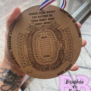 May include: A round, wooden ornament with a stadium seating chart etched on it. The ornament has the text "Where else would you rather be than right here right now?" and "Destiny is calling."