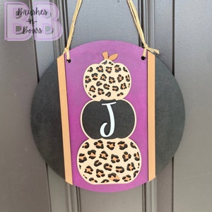 May include: A round wooden door hanger with a purple background and three stacked pumpkins. The pumpkins are decorated with a leopard print pattern and the middle pumpkin has a white letter "J" on it. The door hanger has a brown rope for hanging.