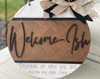 May include: Round wooden sign with a white background and a brown chevron pattern. The sign says "Welcome-Ish" in black script lettering. Below the text, it reads "Depends on who you are and how long you're staying." The sign is decorated with a black and beige burlap bow.