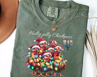 May include: Light green Comfort Colors t-shirt featuring a graphic of six chickens wearing Santa hats and Christmas lights, taking a selfie with the text "Holly Jolly Chickmas" and "Say Chicks".