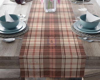 May include: A brown plaid table runner with a mix of light brown, beige, and red-brown tones. The runner is placed on a wooden table set for a meal, with plates, bowls, and wine glasses. A vase of pink flowers sits in the center.
