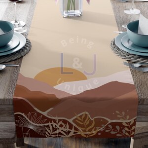 May include: A rectangular table runner featuring a printed landscape design with layered hills in shades of beige, terracotta, and brown. A large yellow sun sits above the hills, with white line-art botanical leaves at the bottom. The text Being L&J Unique is centered.