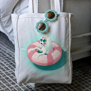 May include: White canvas tote bag featuring a graphic of a white poodle wearing sunglasses and floating in a pink and white striped inner tube. Light blue flower-shaped sunglasses are clipped to the top of the bag.