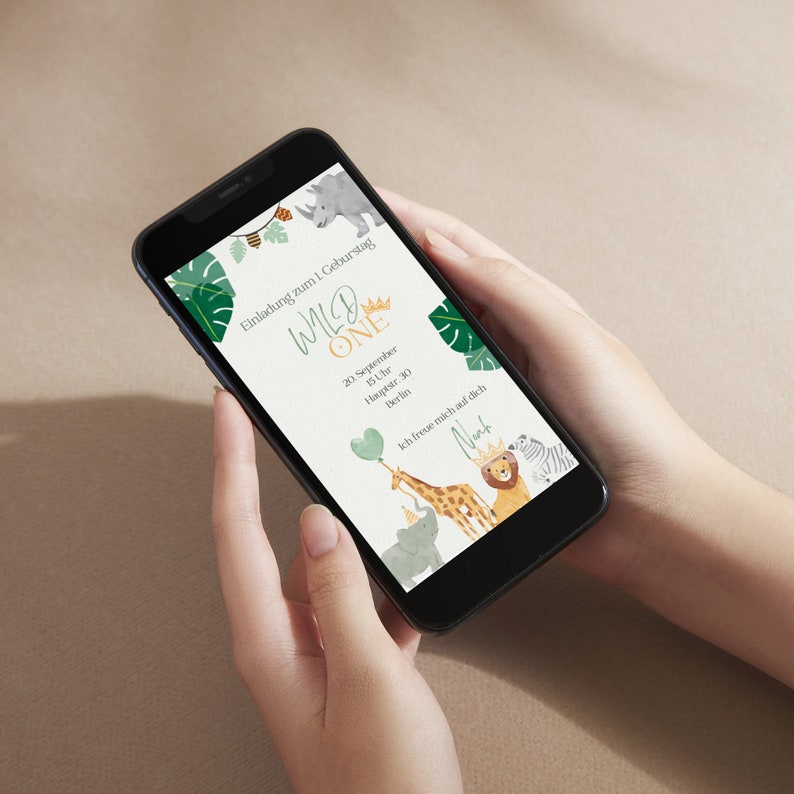 May include: A smartphone displaying a digital invitation for a first birthday party with the text "Wild One" in green and gold lettering. The invitation features a jungle theme with illustrations of animals like a giraffe, lion, zebra, and elephant.