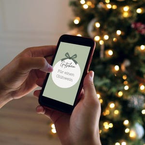 Pu&ograve; includere: Uno smartphone che mostra un buono regalo digitale per un bicchiere di vin brul&egrave;. Il buono regalo &egrave; progettato per assomigliare a un ornamento natalizio con un nastro verde.