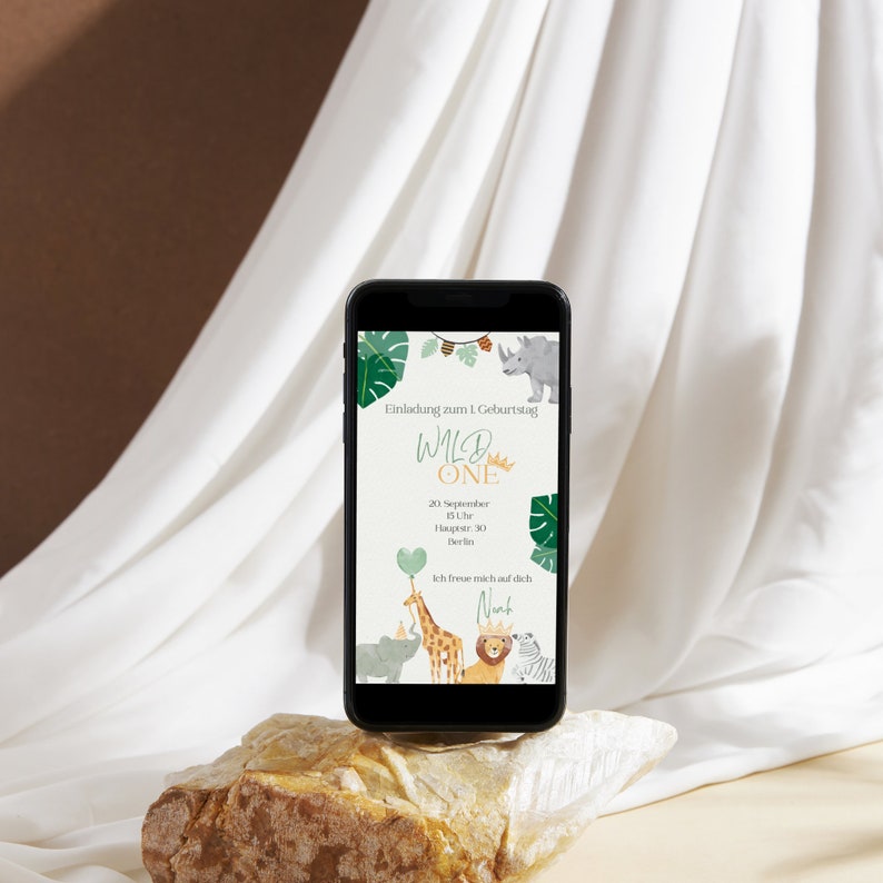 May include: A smartphone displaying a digital invitation for a first birthday party with a jungle theme. The invitation features a green, yellow, and brown color scheme with a rhino, giraffe, lion, and zebra. The text on the invitation reads "Wild One" and includes the date, time, and location of the party. The invitation also includes the name "Nant".