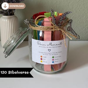 Pode incluir: Um frasco de vidro transparente cheio de tiras de papel coloridas, amarrado com barbante e coberto com lavanda seca. O frasco tem um rótulo com texto em alemão e um símbolo de coração. As palavras "120 Bibelverse" estão na parte inferior.