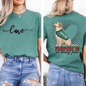 May include: Teal t-shirt with the word "love" in cursive and a paw print design. The back of the shirt features a golden retriever wearing a service dog vest, with the text "Purpose in every paw."