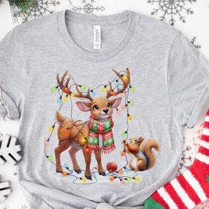 May include: A gray t-shirt with a Christmas-themed design featuring a cartoon deer wearing a scarf and a squirrel hanging from Christmas lights. The shirt is on a white background with snowflakes.
