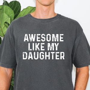 May include: A charcoal gray short-sleeved t-shirt featuring the white distressed-style text "AWESOME LIKE MY DAUGHTER" printed across the chest. The garment has a crew neckline and a relaxed fit, showcasing a solid dark color throughout the fabric.