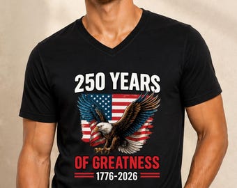 May include: A black V-neck t-shirt featuring a graphic of a bald eagle in front of an American flag. The text reads 250 YEARS OF GREATNESS 1776-2026 in white and red lettering. The design is centred on the chest of the short-sleeved garment.