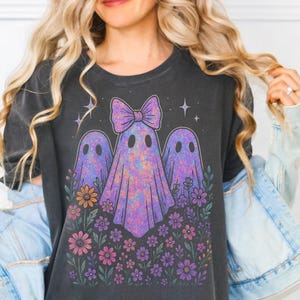 May include: Dark gray t-shirt featuring a whimsical design of three ghosts with floral accents. The ghosts have a purple tie-dye pattern and are adorned with a bow. The design includes flowers in shades of pink, purple, and orange, with stars in the background.