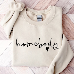May include: A beige sweatshirt with the text "homebody" in black script with a small black heart.
