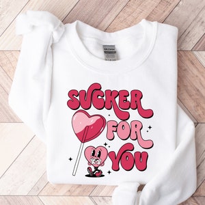 K&ouml;nnte beinhalten: Wei&szlig;er Pullover mit einem pinkfarbenen und roten Grafikdesign, auf dem "SUCKER FOR YOU" steht, mit einem herzf&ouml;rmigen Lutscher und einer Comic-Herzfigur.