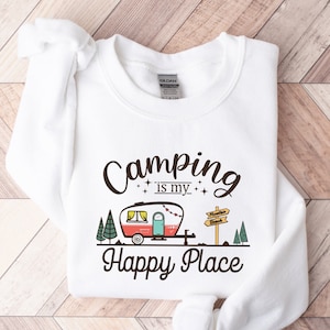 May include: White sweatshirt with the text "Camping is my happy place" in black lettering. A cartoon image of a red and white camper with a green tree on either side of it is below the text. A sign with the text "Adventure awaits" is above the camper.