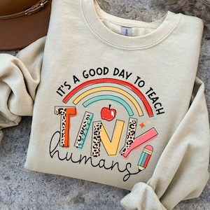 May include: A beige sweatshirt with a rainbow and the text "It's a good day to teach tiny humans". The text is surrounded by a rainbow with a red apple and a pencil.