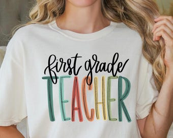May include: A cream-coloured t-shirt featuring the text "first grade TEACHER" in black and colourful letters. The word "TEACHER" is in green, red, yellow, and blue. The shirt has short sleeves and a relaxed fit.