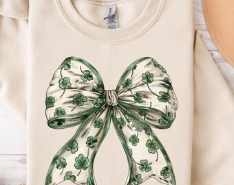 May include: A cream-colored sweatshirt with a large green shamrock bow on the front. The bow is made of a white ribbon with green shamrocks printed on it.