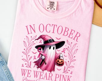 May include: Pink t-shirt with the text "IN OCTOBER WE WEAR PINK Breast Cancer Awareness". The shirt features a ghost wearing a witch hat and holding a pink ribbon and a jack-o'-lantern. The shirt is folded on a white surface.