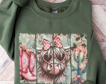 May include: A green sweatshirt with a graphic print of a Highland cow wearing pink heart-shaped sunglasses and a pink bow. The cow is blowing a bubble and there are pink cowboy boots and a cactus with pink hearts on either side of the cow.