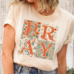 May include: A beige t-shirt with a floral print design that spells out the word "PRAY" in orange letters.