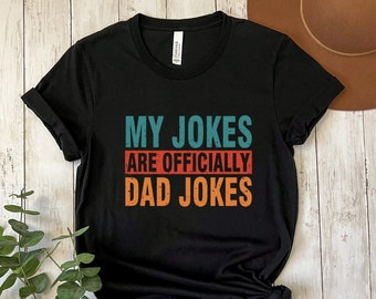 Può includere: Maglietta nera con il testo "My Jokes Are Officially Dad Jokes" in uno schema di colori retrò.