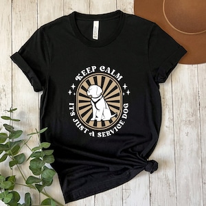 Peut inclure: T-shirt noir avec un motif graphique blanc repr&eacute;sentant un chien portant un gilet de chien d'assistance. Le motif graphique comprend le texte "Keep Calm It's Just a Service Dog" dans un style r&eacute;tro.