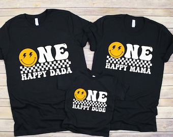 May include: Three black t-shirts with a white checkerboard pattern and a yellow smiley face with lightning bolts. The text "ONE" is in white with a retro font. The shirts say "Happy Dada", "Happy Mama", and "Happy Dude".