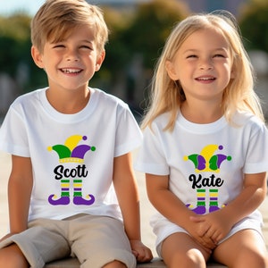 May include: Two children wearing white t-shirts with a Mardi Gras jester design and their names, Scott and Kate, printed on the shirts.