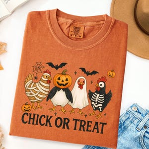 May include: Orange t-shirt with a Halloween-themed graphic. The design features four chickens dressed as a mummy, a pumpkin, a ghost, and a skeleton, with bats and pumpkins. The text "CHICK OR TREAT" is printed below the chickens.