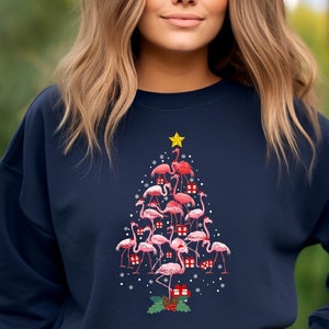 May include: A navy blue sweatshirt with a Christmas tree design featuring pink flamingos and red gift boxes. The tree is topped with a yellow star.