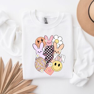May include: White sweatshirt featuring a graphic design with pink and orange bunnies, a chequered lightning bolt, a smiley face, a flower, and a heart. The design is in a retro style.