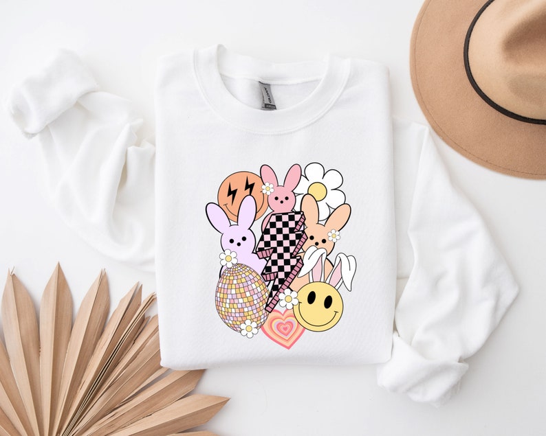 May include: White sweatshirt featuring a graphic design with pink and orange bunnies, a chequered lightning bolt, a smiley face, a flower, and a heart. The design is in a retro style.