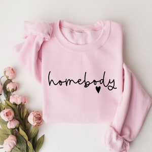 May include: A pink sweatshirt with the text "homebody" in black script with a small black heart.