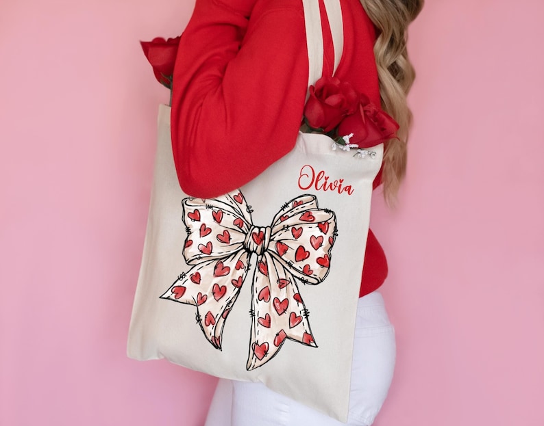 K&ouml;nnte beinhalten: Eine wei&szlig;e Canvas-Tragetasche mit einer roten Schleife und einem Herz-Druck. Die Tasche ist mit dem Namen "Olivia" personalisiert, der auf der Vorderseite aufgedruckt ist.