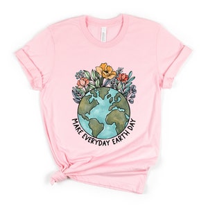 May include: A light pink t-shirt featuring a watercolor illustration of the Earth with flowers and the text "MAKE EVERYDAY EARTH DAY" in black. The shirt is made of a soft material and has rolled-up sleeves.