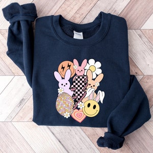 May include: A navy blue sweatshirt featuring a graphic design of pastel pink and orange bunnies, a yellow smiley face, a checkered pattern, and a daisy. The graphic is centred on the sweatshirt.