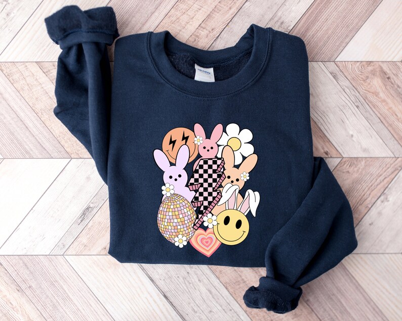May include: A navy blue sweatshirt featuring a graphic design of pastel pink and orange bunnies, a yellow smiley face, a checkered pattern, and a daisy. The graphic is centred on the sweatshirt.