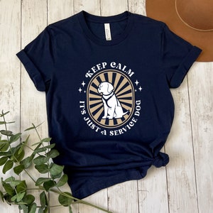 Peut inclure: T-shirt bleu marine avec un graphique blanc d'un chien dans un cercle avec le texte "Keep Calm It's Just a Service Dog".