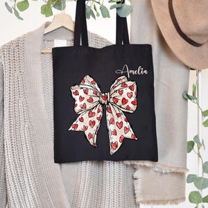 K&ouml;nnte beinhalten: Schwarze Tragetasche mit einem gro&szlig;en roten und wei&szlig;en Bogen mit Herzen darauf gedruckt. Die Tasche tr&auml;gt den Namen Amelia in wei&szlig;.