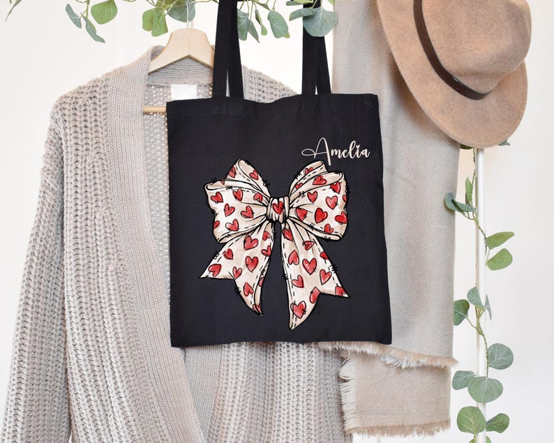 K&ouml;nnte beinhalten: Schwarze Tragetasche mit einem gro&szlig;en roten und wei&szlig;en Bogen mit Herzen darauf gedruckt. Die Tasche tr&auml;gt den Namen Amelia in wei&szlig;.