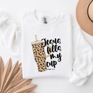 May include: White sweatshirt with a black and brown leopard print tumbler and the text "Jesus fills my cup" with the scripture reference "Psalm 23:5".