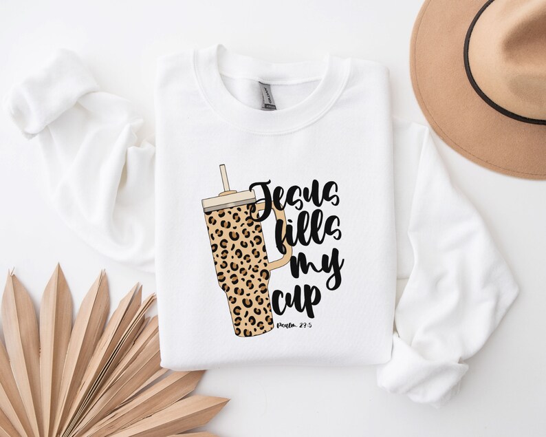 May include: White sweatshirt with a black and brown leopard print tumbler and the text "Jesus fills my cup" with the scripture reference "Psalm 23:5".