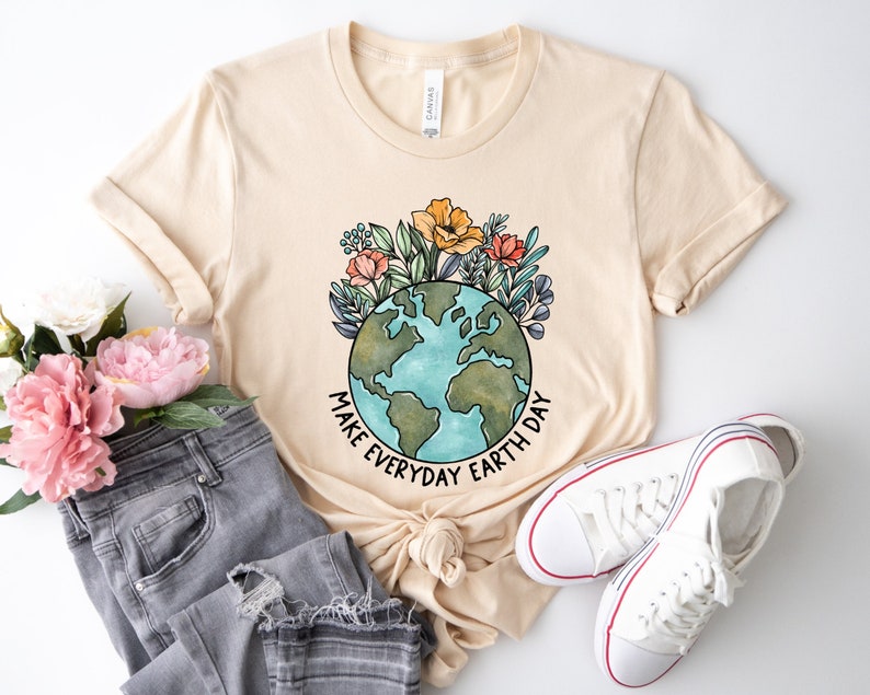 May include: A cream-colored t-shirt featuring a watercolor illustration of the Earth surrounded by colorful flowers. The text "MAKE EVERYDAY EARTH DAY" is printed in black. The shirt is styled with gray jeans and white sneakers.