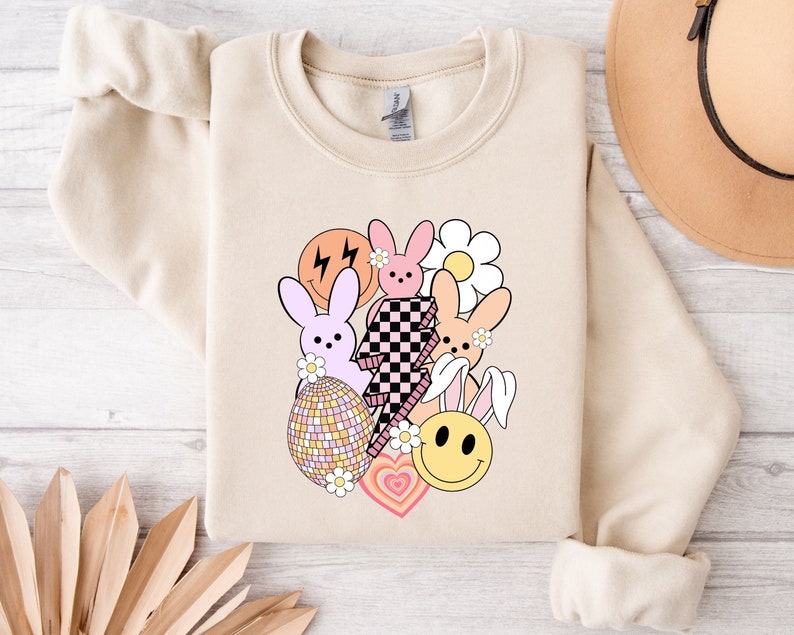 May include: A beige sweatshirt with a graphic design featuring a pink, purple, and orange bunny rabbit, a chequered pattern, a smiley face, a flower, and a heart. The design is in a retro style.