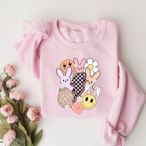 May include: Pink sweatshirt featuring a graphic design of cartoon bunnies, hearts, and a smiley face. The design is in pastel colours and includes a checkered pattern.