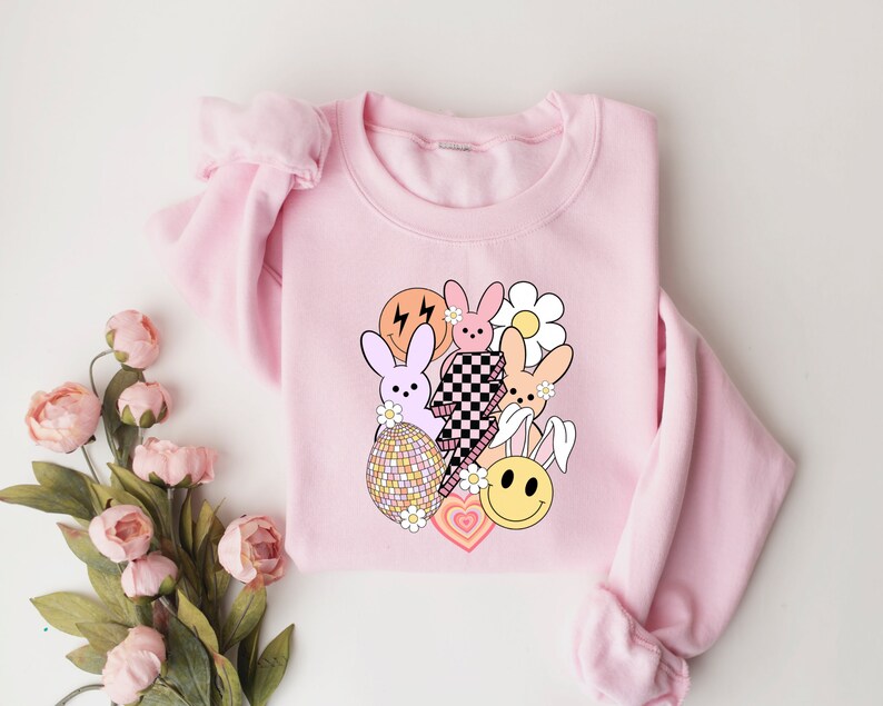 May include: Pink sweatshirt featuring a graphic design of cartoon bunnies, hearts, and a smiley face. The design is in pastel colours and includes a checkered pattern.