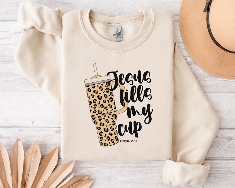 May include: A cream-colored sweatshirt with a leopard print tumbler and the text "Jesus fills my cup, Psalm 23:5".