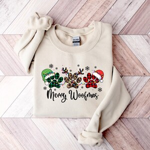 May include: A cream-coloured sweatshirt with a Christmas-themed design featuring three paw prints wearing Santa hats. The paw prints are decorated with green and red plaid, leopard print, and green and white plaid. The text "Merry Woofmas" is printed below the paw prints.